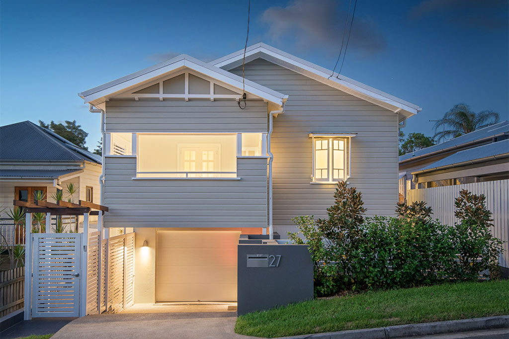 Evening view of a renovated Queenslander-style home on a narrow lot in Red Hill, Brisbane. The design features a raised entry with integrated garage, white timber cladding, soft lighting, and clean landscaping. The layout creates greater privacy and street appeal.