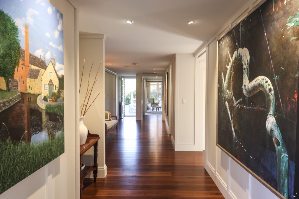 Interior hallway from the Gregors Creek Custom New Home Design, showing rich timber flooring, large-scale nature-inspired artwork, and long views through to a sunlit living space and greenery beyond. The space feels calm and grounded.