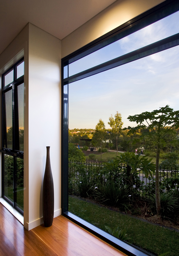 Interior view of a modern Brisbane home with full-height corner windows offering uninterrupted views to a green garden and open sky. Timber floors and minimalist decor enhance the natural connection.