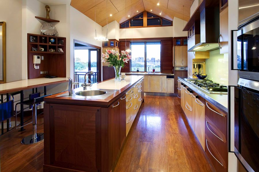 A custom-designed kitchen in a Brisbane home featuring rich timber cabinetry, polished timber flooring, and a large island bench with a built-in sink and floral centrepiece. The space includes integrated appliances, a long cooktop with overhead rangehood, and a light-filled dining nook with built-in shelving. A vaulted timber ceiling and large windows create a warm, open atmosphere that supports family living and entertaining.