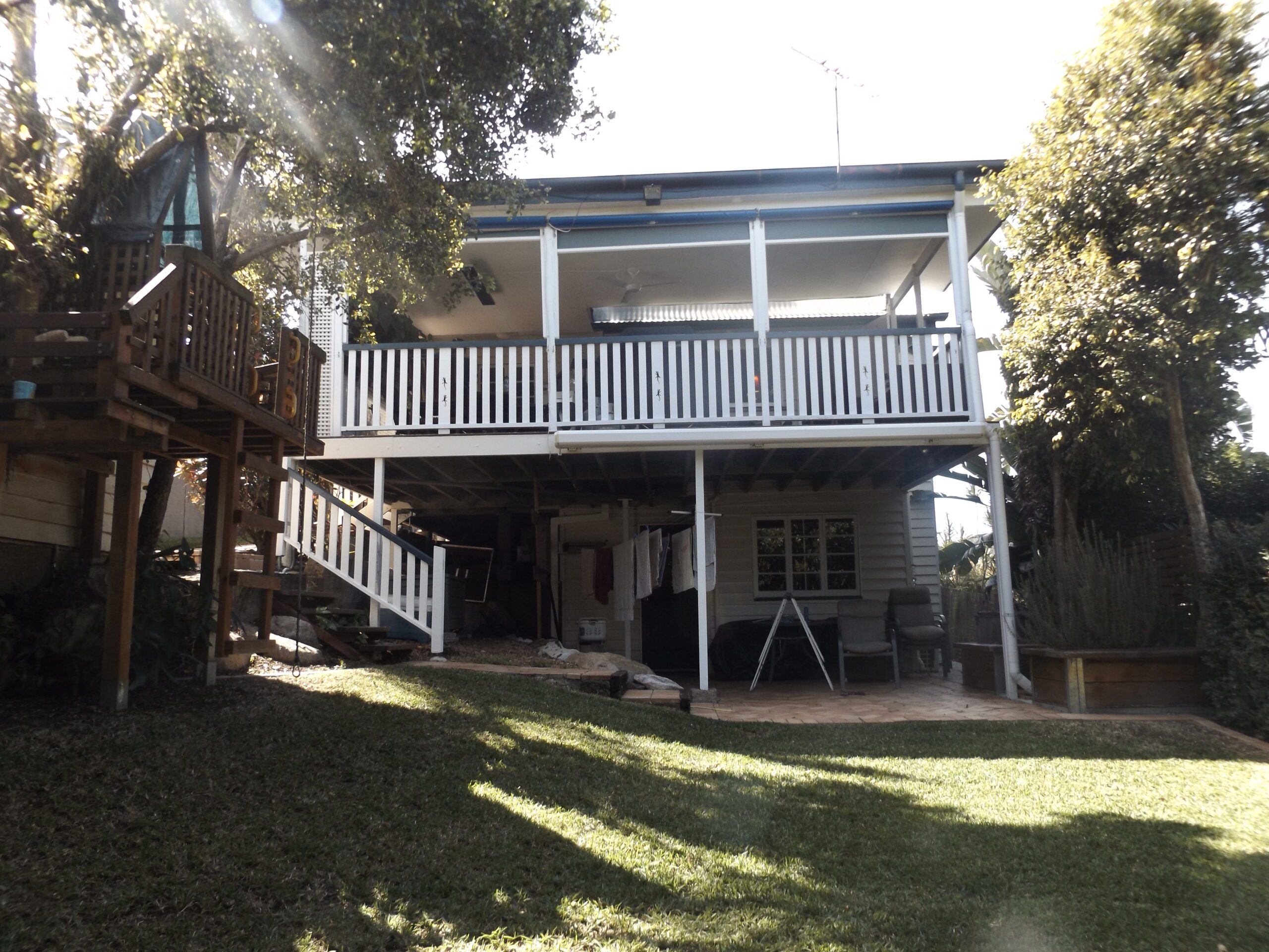 Pre-renovation Queenslander in Taringa with two-story rear elevation, upper-level veranda, exposed ground floor, and sloped backyard with established trees