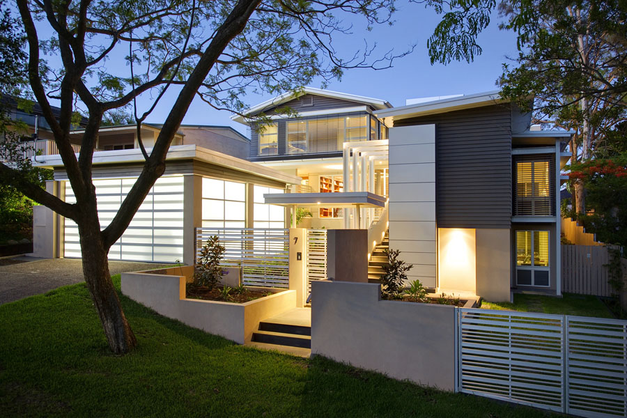 Evening view of a modern renovated home in Indooroopilly featuring layered levels, timber and rendered finishes, horizontal aluminium louvre screens, and a softly illuminated garden and entrance pathway framed by mature trees.
