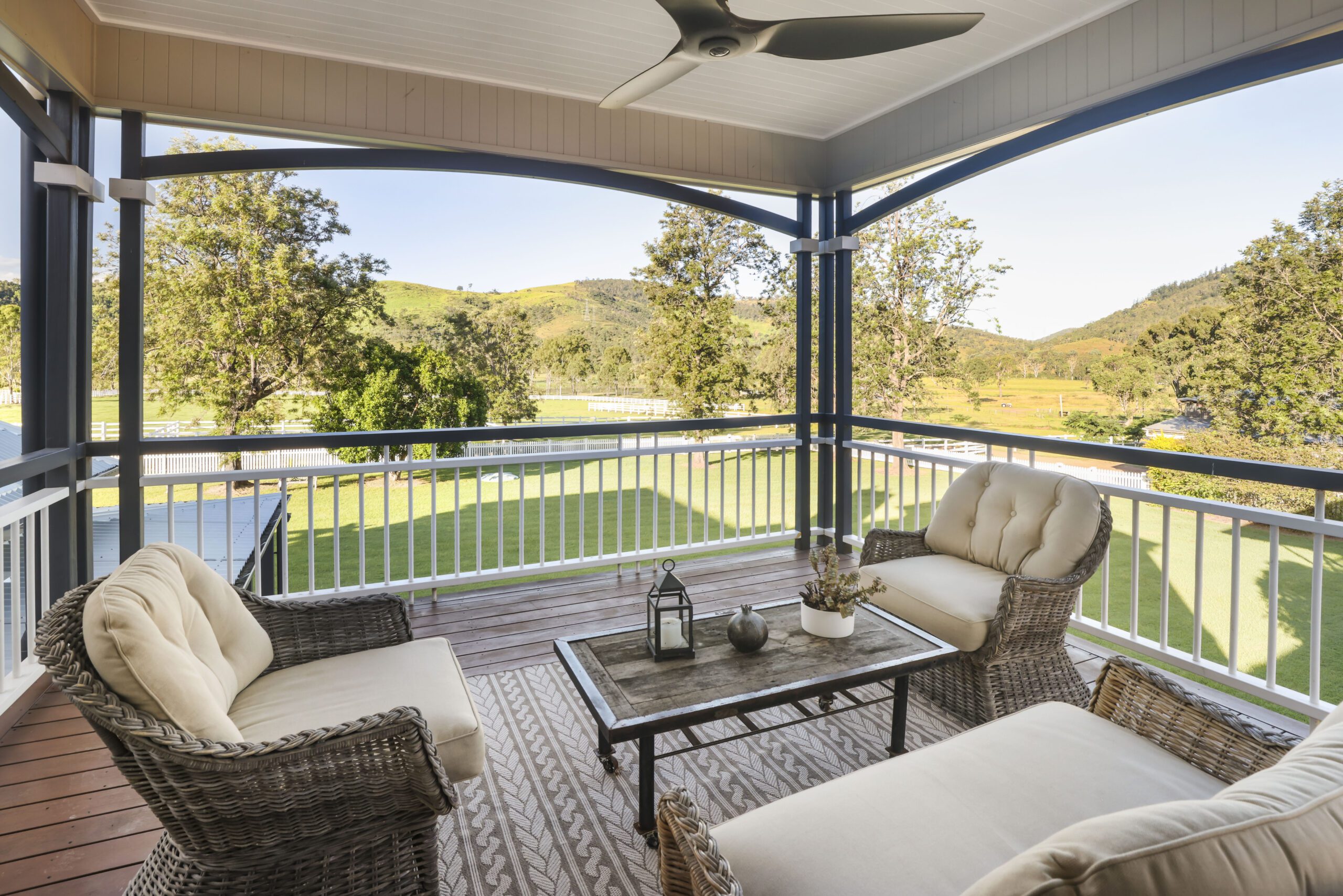 Covered veranda with wicker sofas around a coffee table, white balustrade, ceiling fan and open views across lawns, trees and rolling hills.