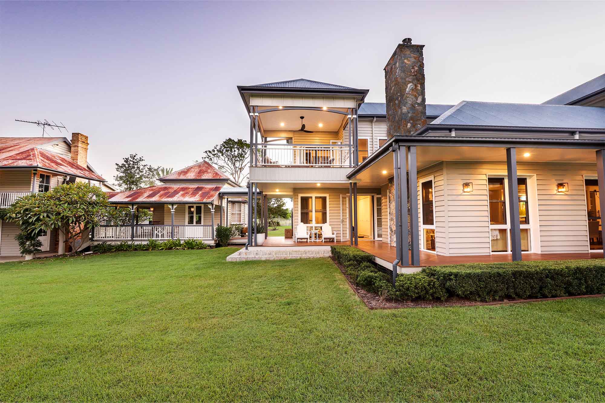 Two-storey modern country-style home designed by a Brisbane architect, featuring a wide lawn, wrap-around verandas, and a combination of stone and timber finishes under a pitched roof.