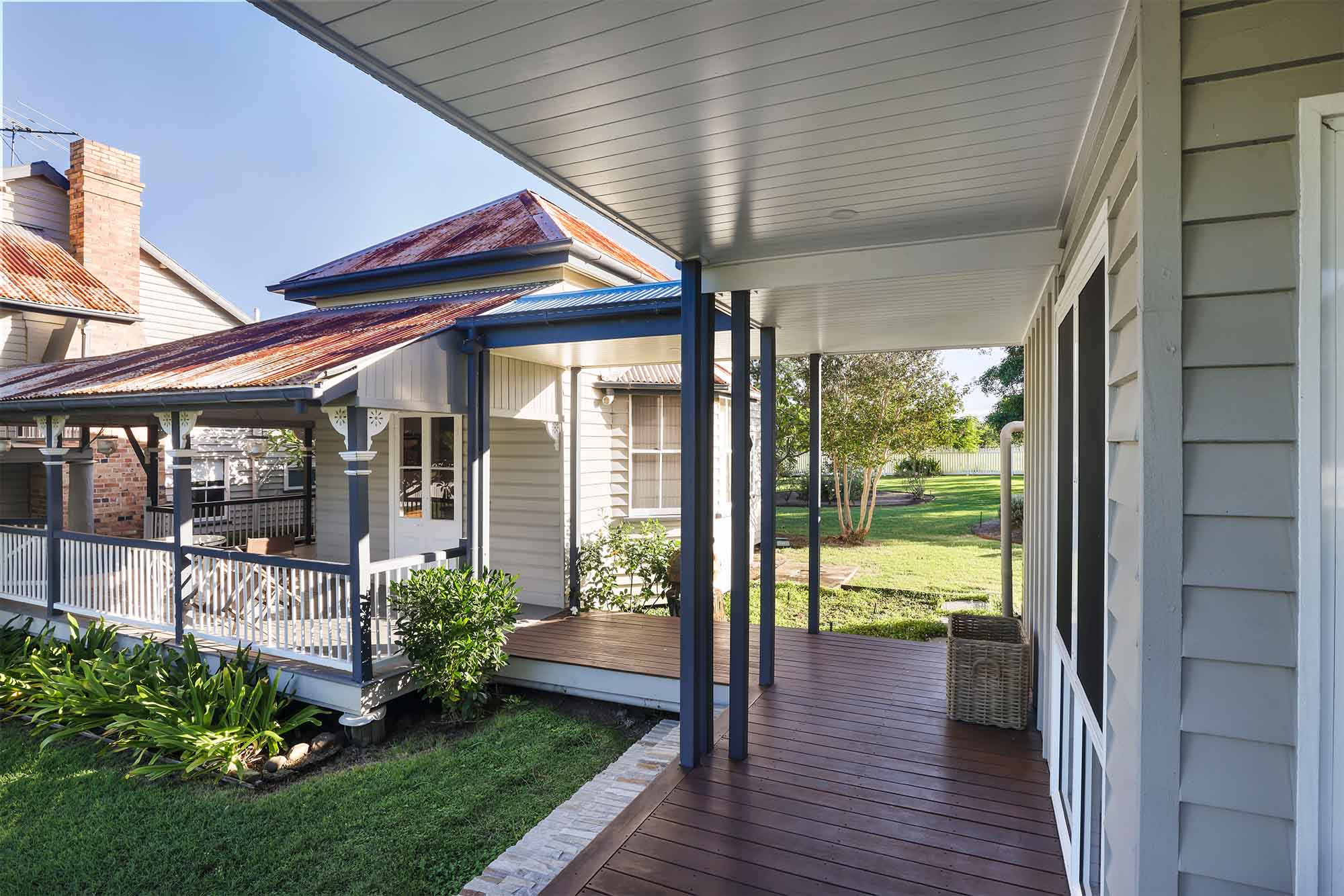 Covered verandah walkway connecting a renovated Queenslander home with a new secondary dwelling. The original house features timber weatherboards, ornate posts, and a corrugated iron roof with areas of natural rust. The new extension continues the timber cladding with fresh paint and a modern deck in rich timber tones. Green lawns, garden beds, and mature trees surround the property, blending the old and new architecture into a cohesive homestead setting.