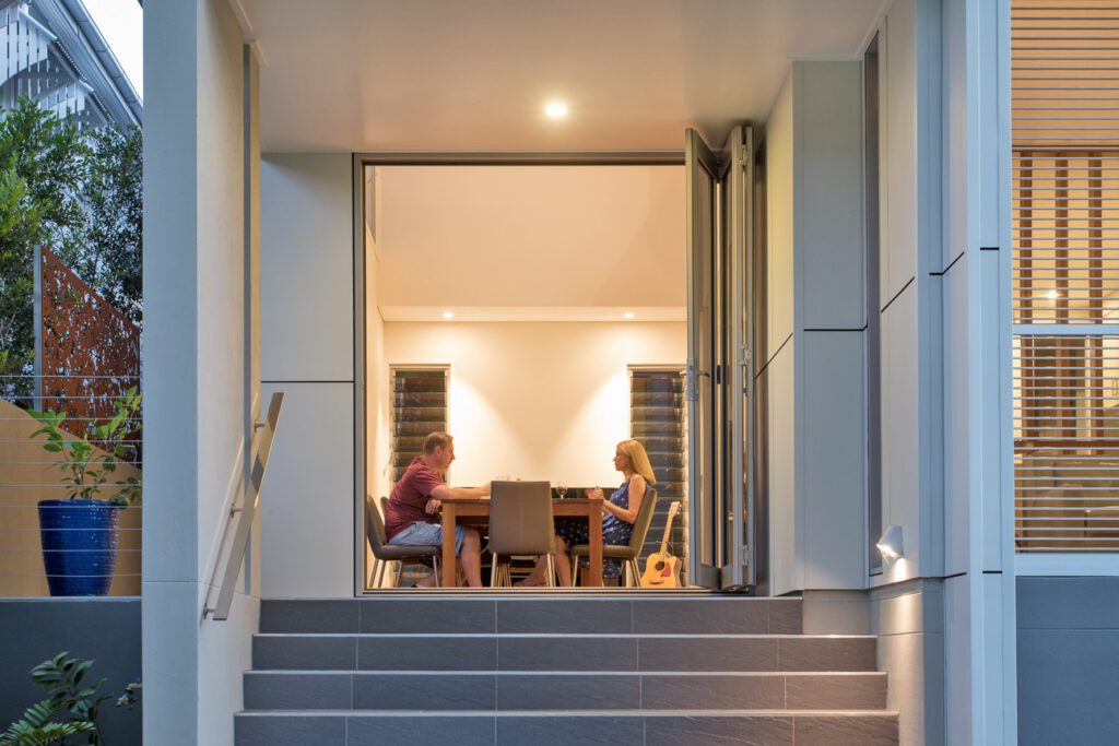 Evening view of a Brisbane home’s dining area with bifold doors fully open, showing a couple enjoying dinner at a timber table, framed by modern cladding and soft lighting.