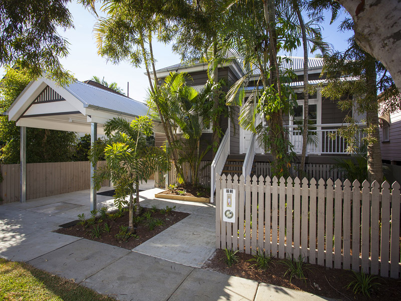 Front of a renovated home with a new white picket fence, landscaped garden, modern carport with gabled roof, and visible welcoming entry stairs, creating strong street appeal.