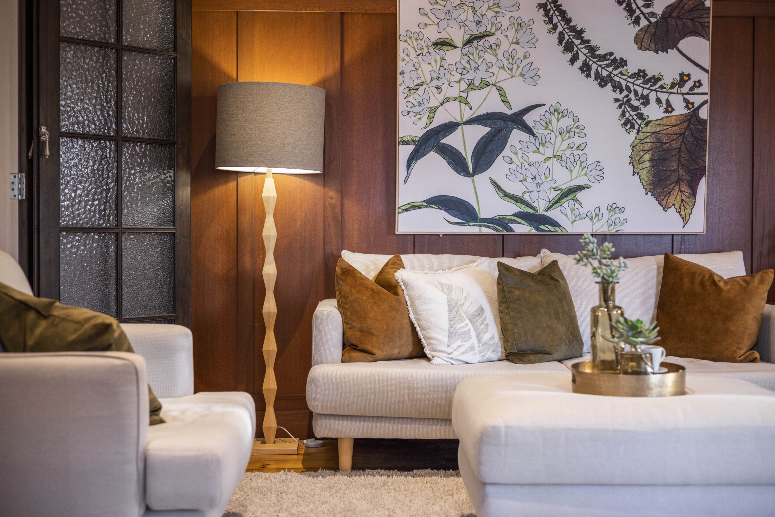 Warm and inviting living room with timber wall panelling, modern cream sofas, soft brown and white cushions, standing floor lamp, and large botanical artwork.