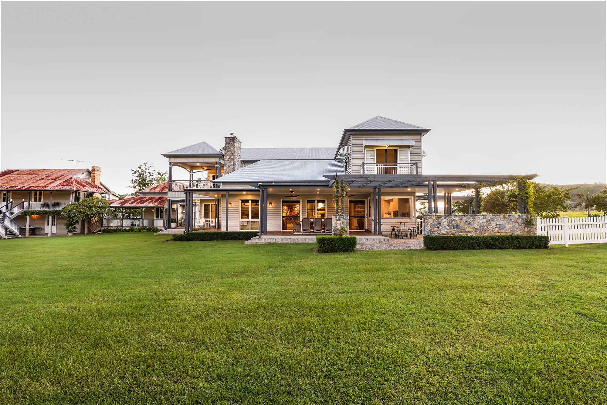 Two-storey country-style home with grey weatherboard cladding, wraparound verandahs, stone chimney, and landscaped lawn in a rural setting at Gregor’s Creek.