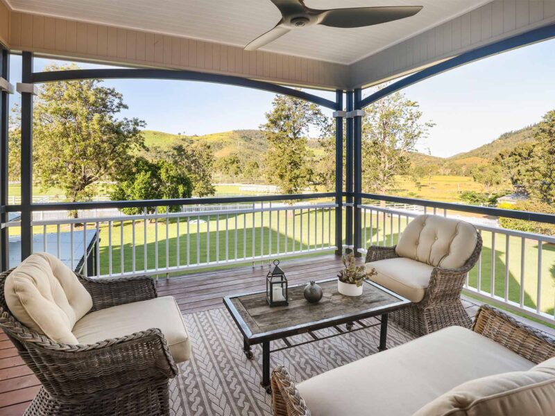 Covered verandah with cushioned wicker chairs and a timber coffee table overlooking green paddocks and rolling hills, framed by white balustrades and a ceiling fan.