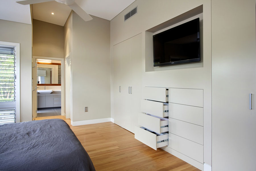 Bedroom with built-in cabinetry featuring integrated wardrobes, a recessed TV niche, and a bank of custom drawers. Timber flooring and louvre windows bring warmth and natural light into the space.