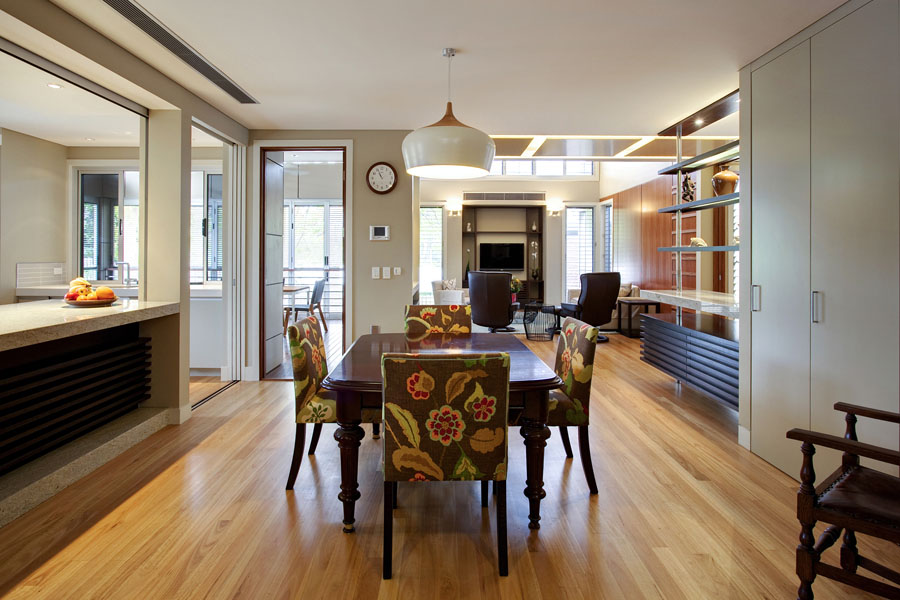 “Open-plan dining and living area with timber flooring and warm neutral tones. A wooden dining table with patterned upholstered chairs sits centrally beneath a modern pendant light. The space connects to the kitchen through a wide servery opening, and the adjoining living room features built-in shelving, a TV unit, and large windows that bring in natural light.